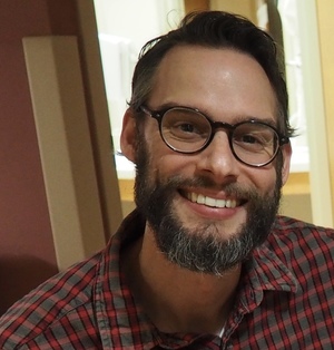 Bearded man with gray-brown hair and glasses, wearing a flannel shirt and smiling.