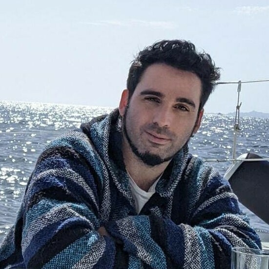 Man with dark hair and beard wearing blue striped hoodie, seated with arms crossed over chest before the sea.