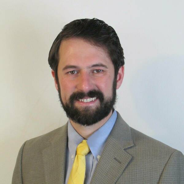 Young man with brown hair and beard wearing blue oxford shirt, yellow tie, and gray suit jacket.