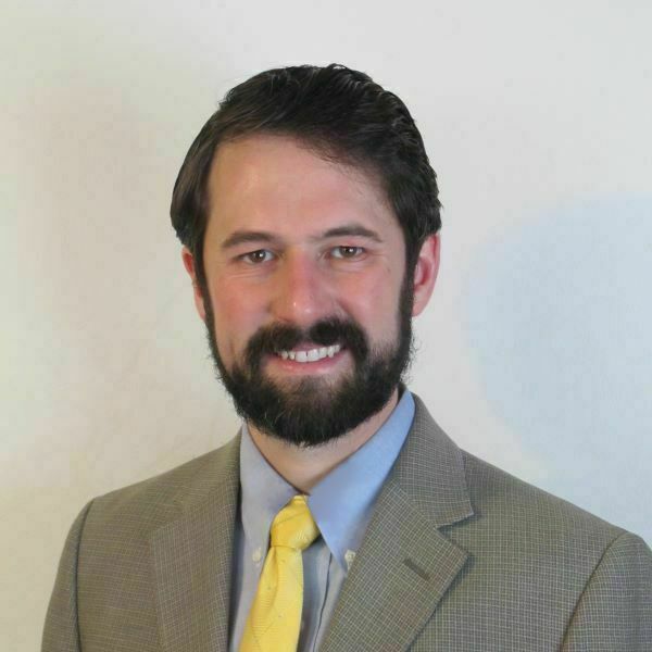 Young man with brown hair and beard wearing blue oxford shirt, yellow tie, and gray suit jacket.