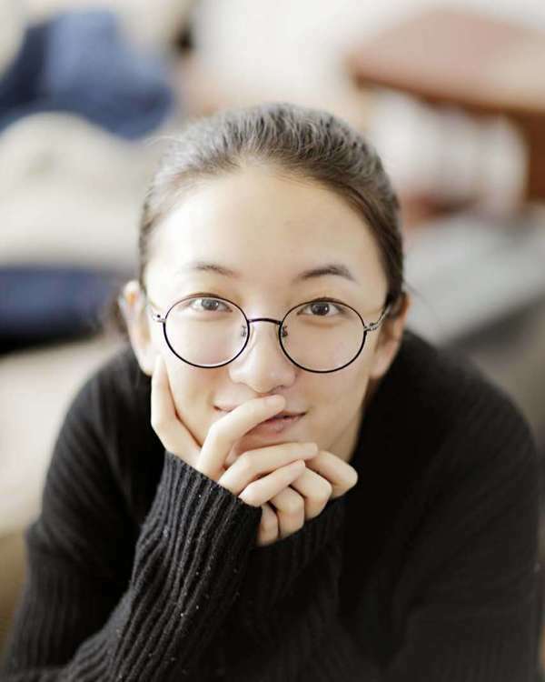 Young woman with round glasses and light brown hair, wearing black sweater, smiling with hand raised to chin.
