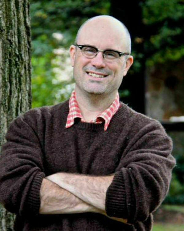 Man with shaved head and horned rimmed glasses, wearing brown sweater and red-and-white checkered shirt, with crossed arms, standing before tree.