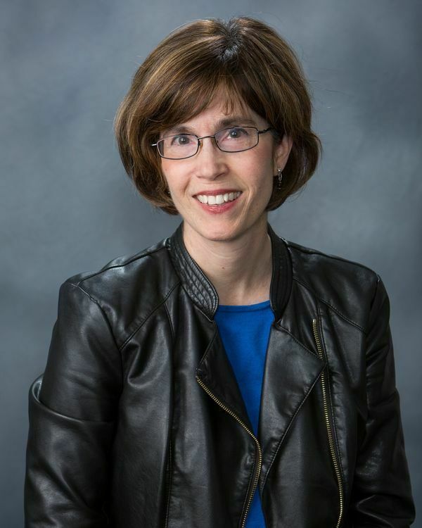 Woman with short brown hair and glasses, wearing black leather jacket and blue blouse, smiling
