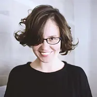 A woman with short, dark wavy hair and black-framed glasses smiles broadly, wearing a black shirt.